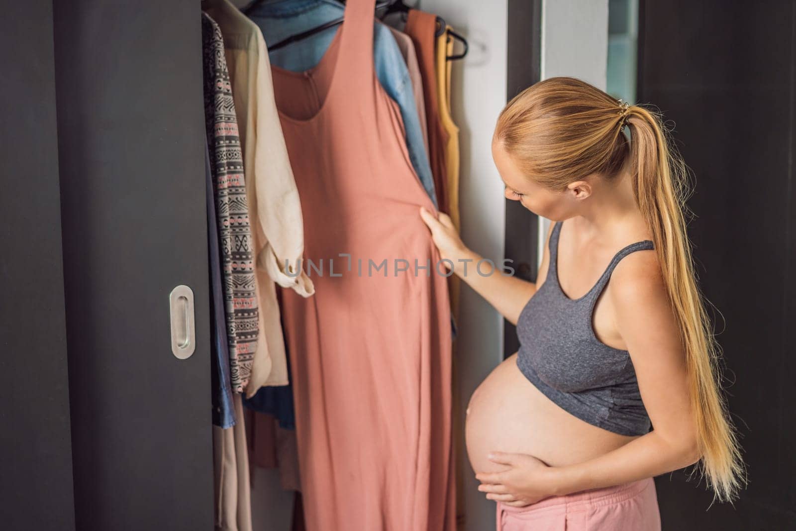 A pregnant woman has nothing to wear. A pregnant woman stands in front of a closet with clothes and does not know what to wear because the clothes do not fit on her.