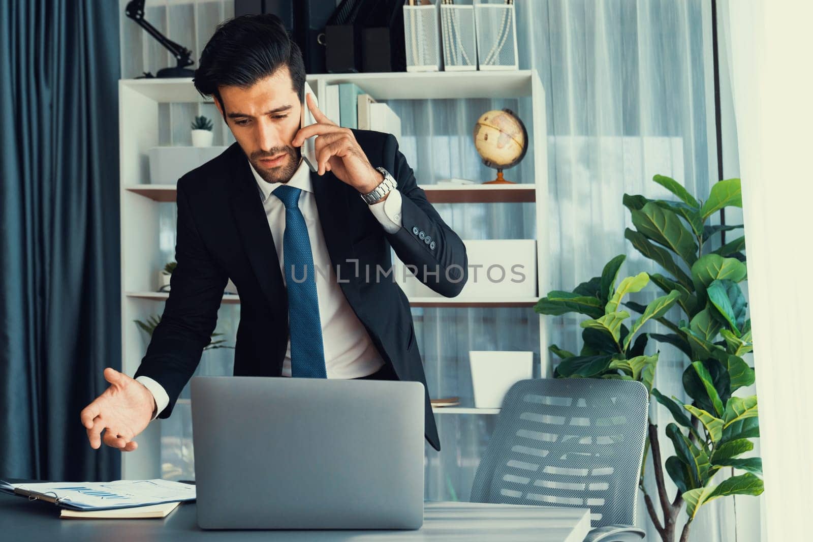 Diligent businessman busy talking on the phone call with clients while working with laptop in his office as concept of modern hardworking office worker lifestyle with mobile phone. Fervent