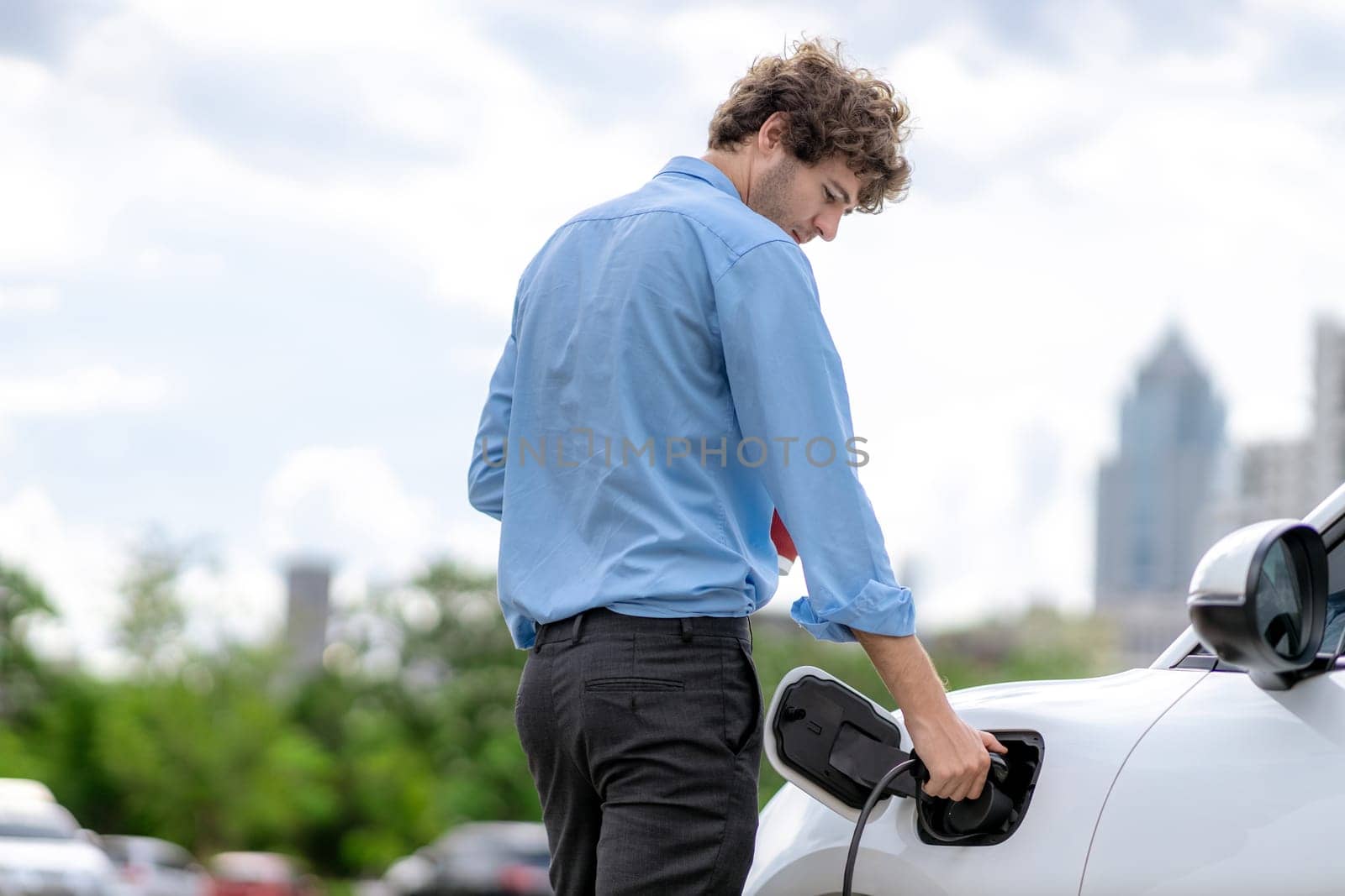 Progressive eco-friendly concept of parking EV car at public electric-powered charging station in city with blur background of businessman leaning on recharging-electric vehicle with coffee.