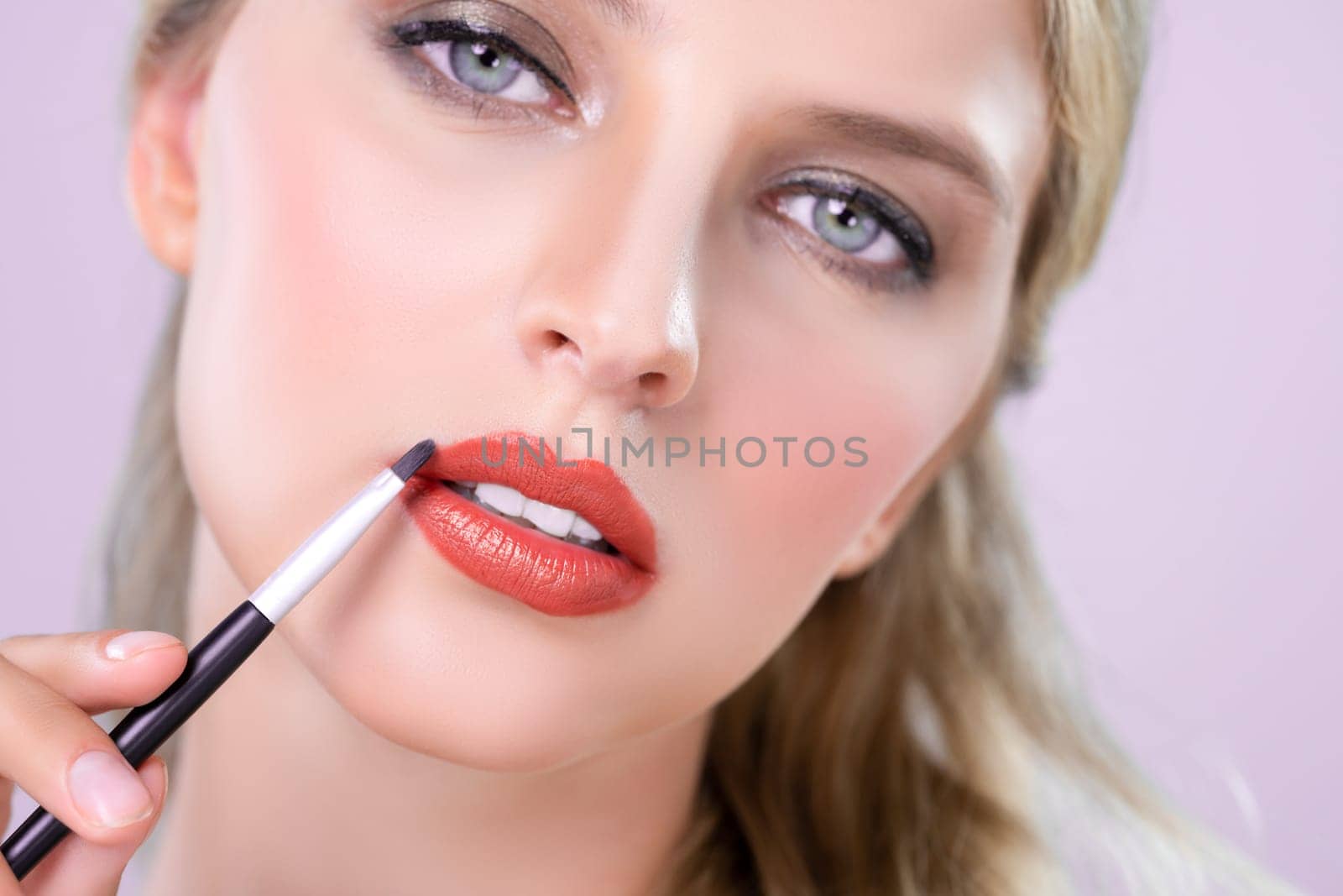 Closeup beautiful young woman with flawless healthy skin and natural makeup putting alluring fashion glossy red lipstick on her lip with lip brush in pink isolated background.