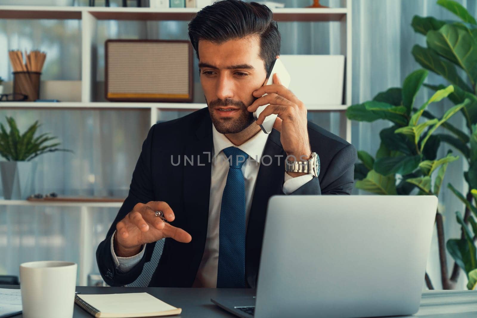 Diligent businessman busy talking on the phone call with clients while working with laptop in his office as concept of modern hardworking office worker lifestyle with mobile phone. Fervent