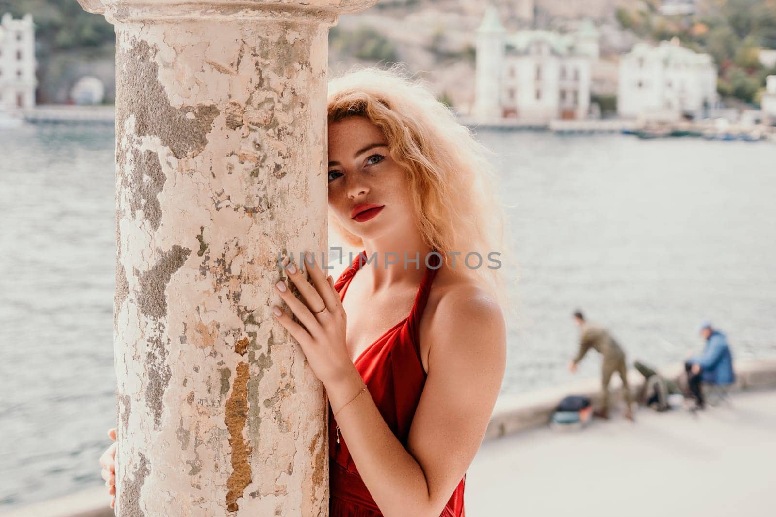 Close up shot of beautiful young caucasian woman with curly blond hair and freckles looking at camera and smiling. Cute woman portrait in a pink long dress posing on a volcanic rock high above the sea