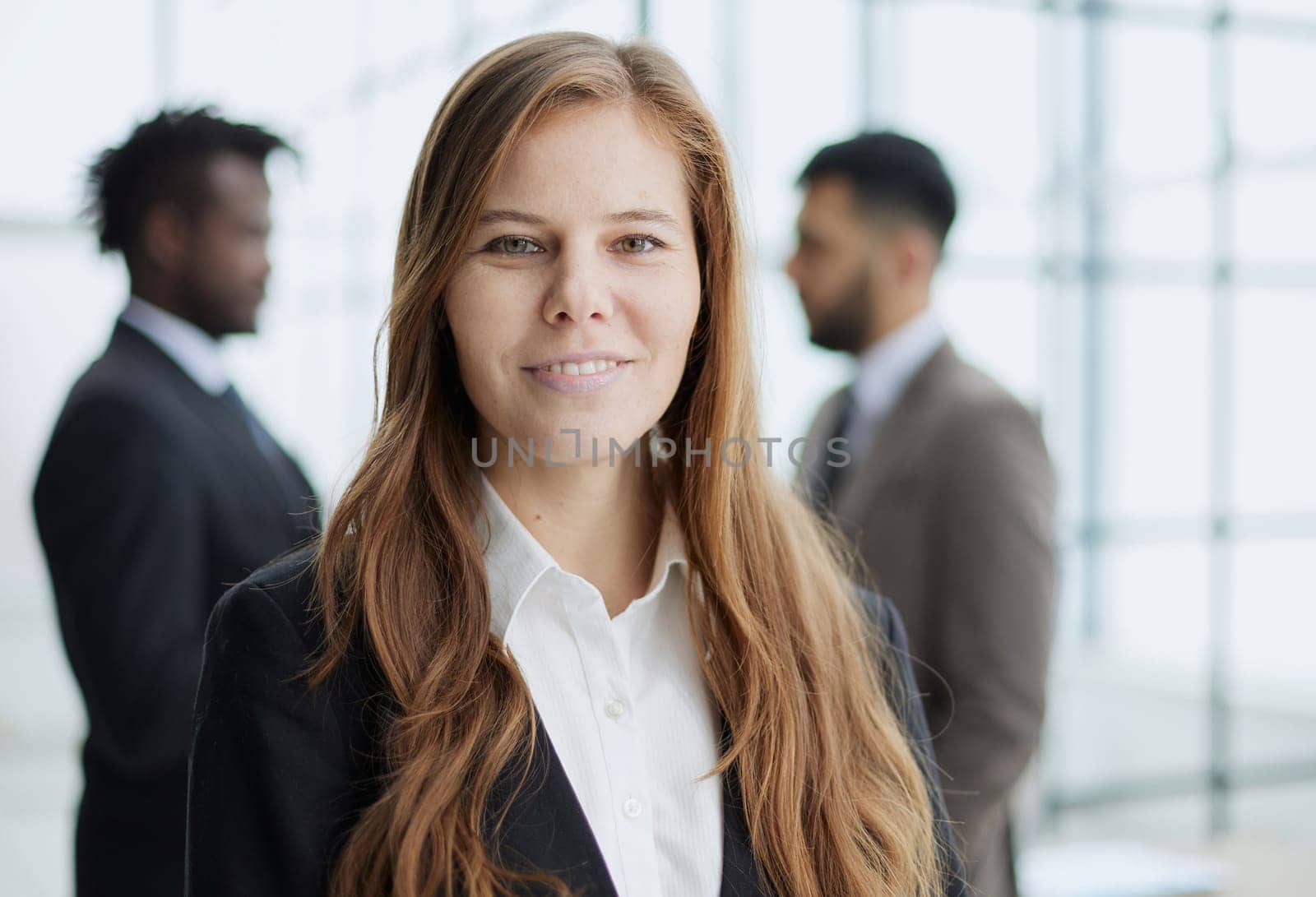 A beautiful business woman stands and looks confidently into the camera against the background of her colleagues