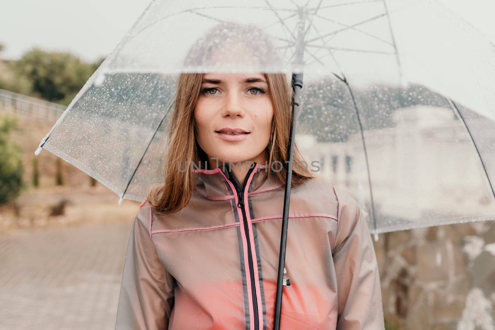 Woman rain umbrella. Happy woman portrait wearing a raincoat with transparent umbrella outdoors on rainy day in park near sea. Girl on the nature on rainy overcast day. by panophotograph