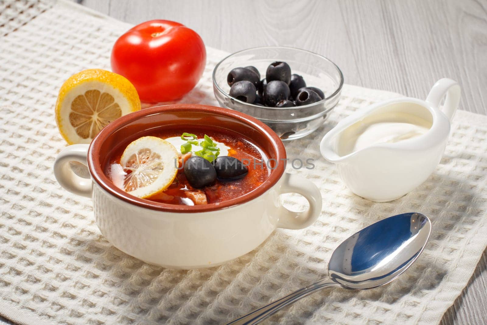Soup saltwort with meat, smoked sausages, potatoes, tomatoes, marinated pickled cucumber, lemon, black olives and sour cream, ceramic soup bowl with ingredients on white kitchen towel.