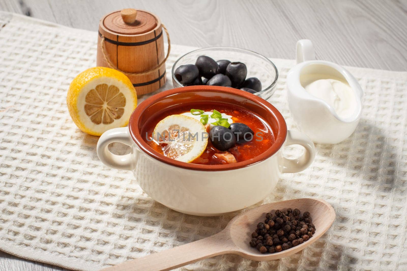 Soup saltwort with meat, sausages, potatoes, tomatoes, marinated pickled cucumber, lemon, black olives and sour cream with ingredients, wooden spoon with peppercorns on kitchen towel.