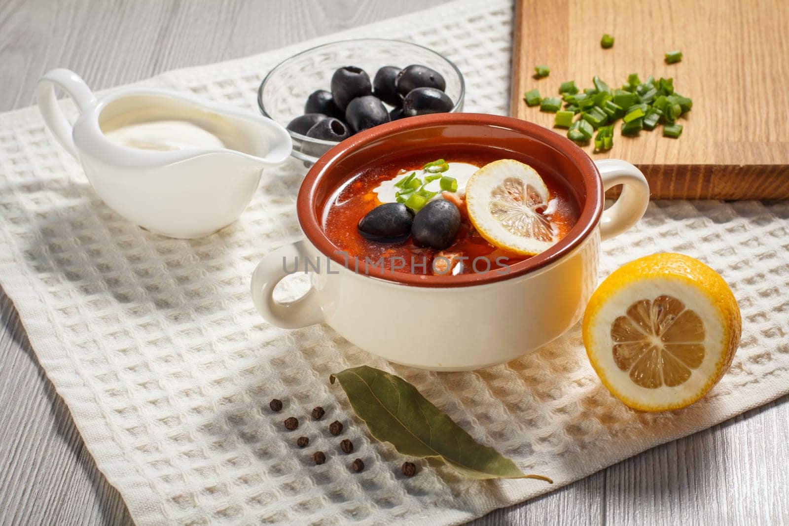 Soup saltwort with meat, smoked sausages, potatoes, tomatoes, marinated pickled cucumber, lemon, black olives and sour cream in ceramic soup bowl with ingredients and peppercorns.
