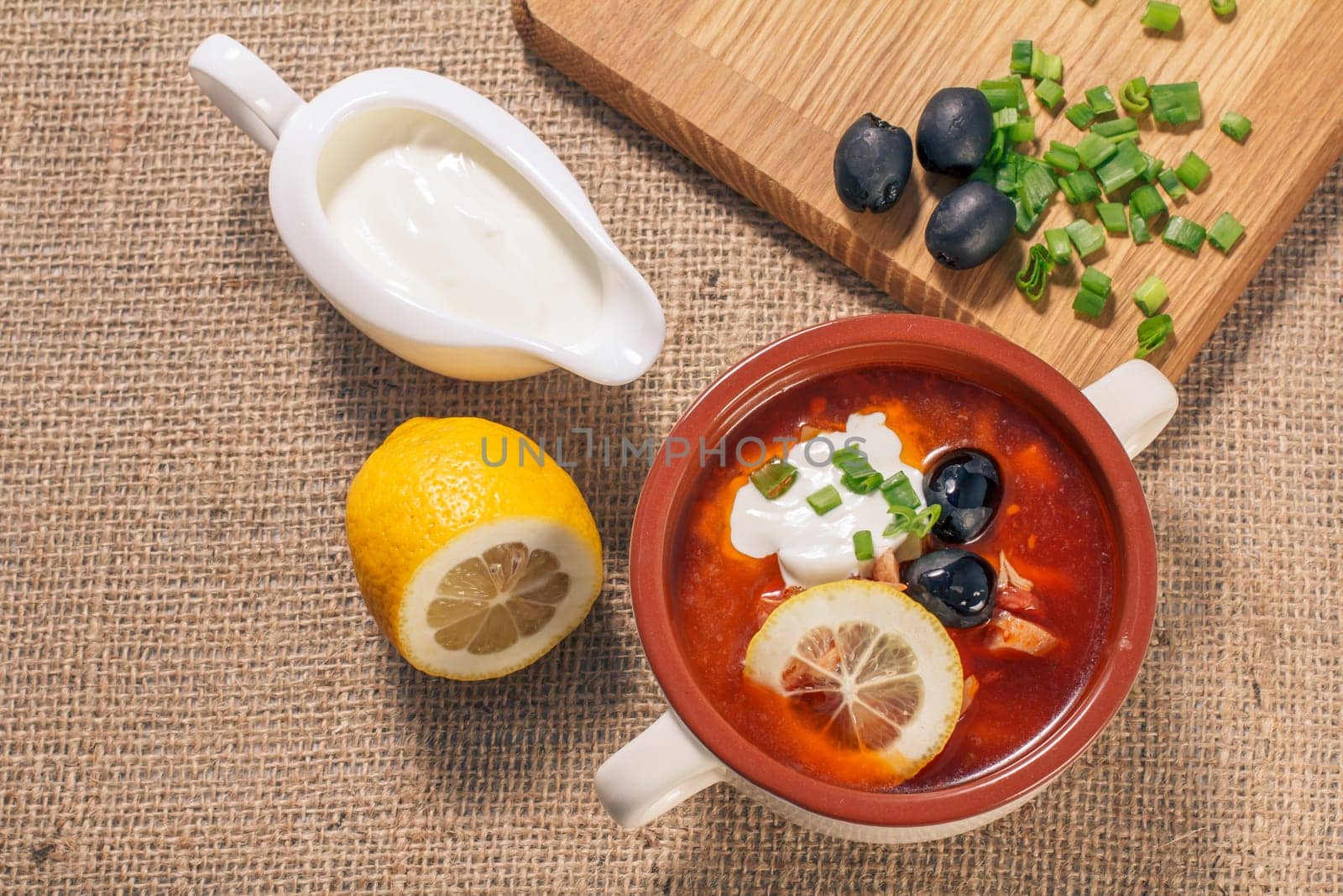 Soup saltwort with meat, smoked sausages, potatoes, tomatoes, marinated pickled cucumber, lemon, black olives and sour cream in ceramic soup bowl with ingredients. Top view