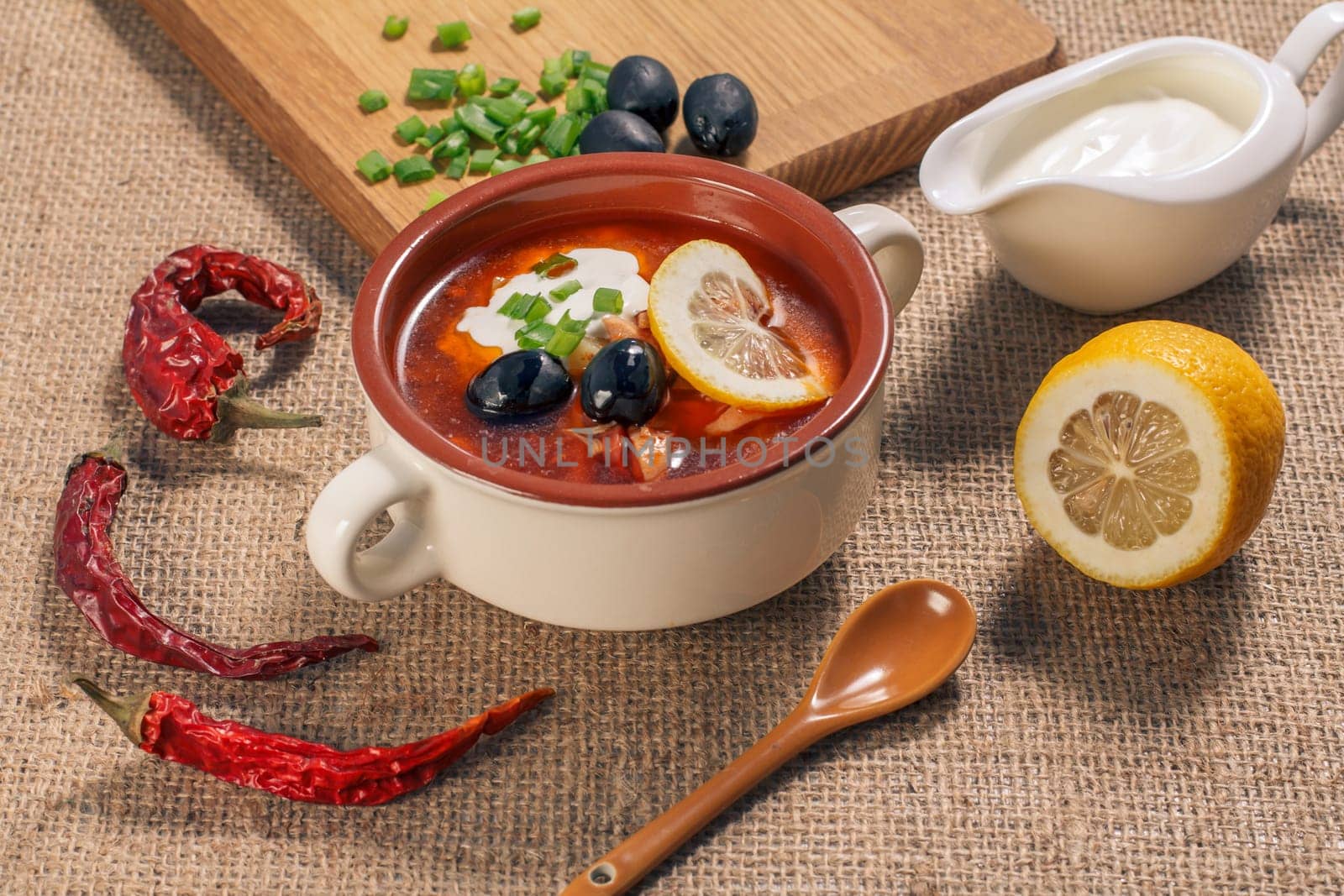 Soup saltwort with meat, smoked sausages, potatoes, tomatoes, marinated pickled cucumber, lemon, black olives and sour cream in ceramic soup bowl with ingredients.