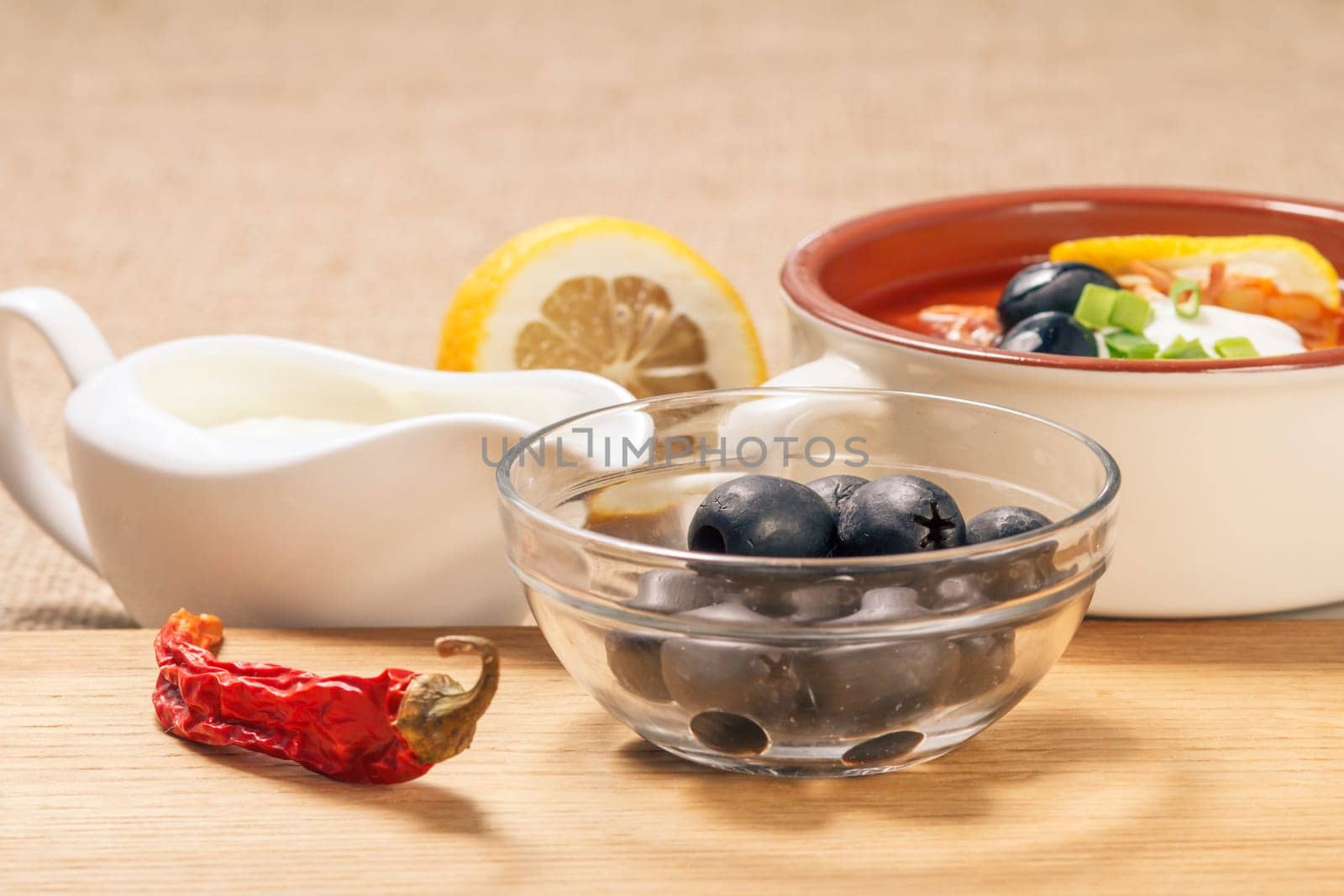 Glass bowl with black olives, dried red pepper on cutting board and ceramic soup bowl with saltwort, sauceboat with sour cream and cut lemon in the background.
