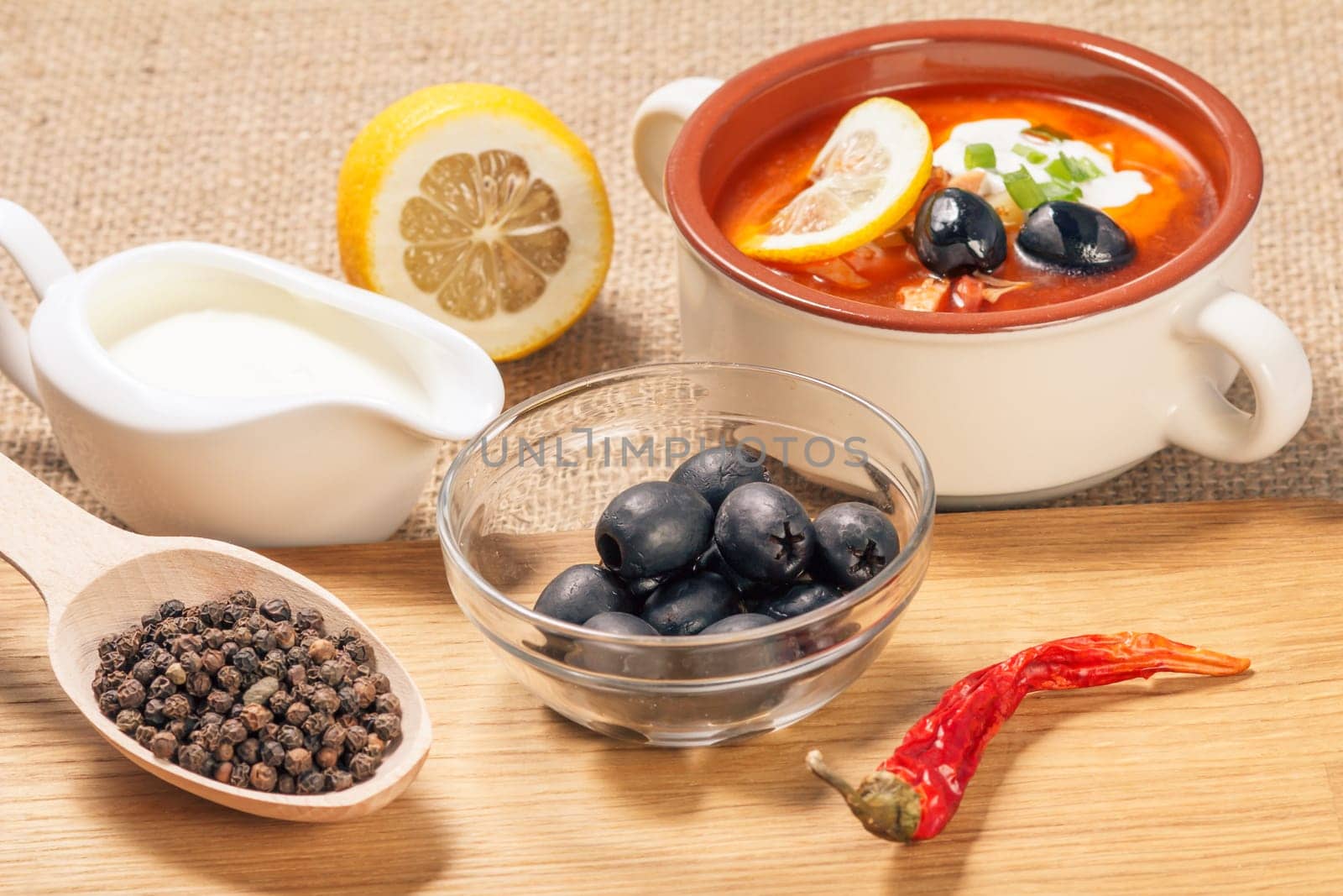 Wooden spoon with black peppercorn, glass bowl with olives, dried red pepper on cutting board and ceramic soup bowl with saltwort, sauceboat with sour cream and cut lemon. Top view.
