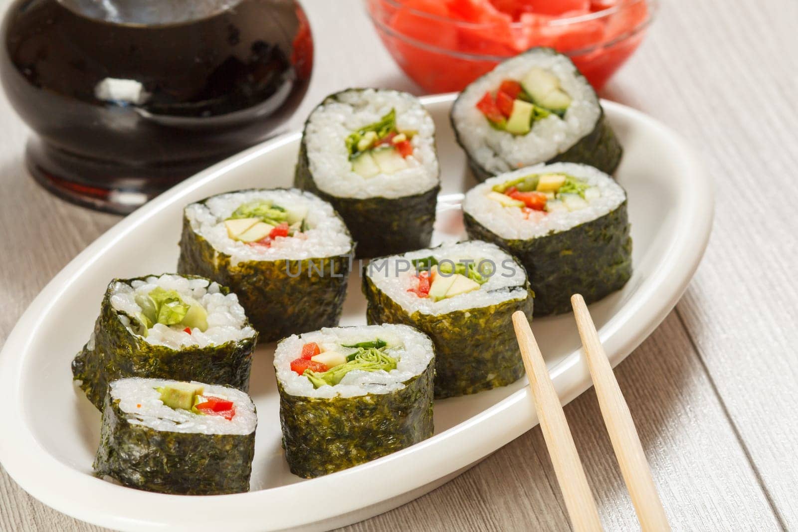 Sushi rolls with rice, pieces of avocado, cucumber, red bell pepper and lettuce leaves on ceramic plate, chopsticks, glass bottle with soy sauce and pickled ginger in a bowl. Vegetarian food.