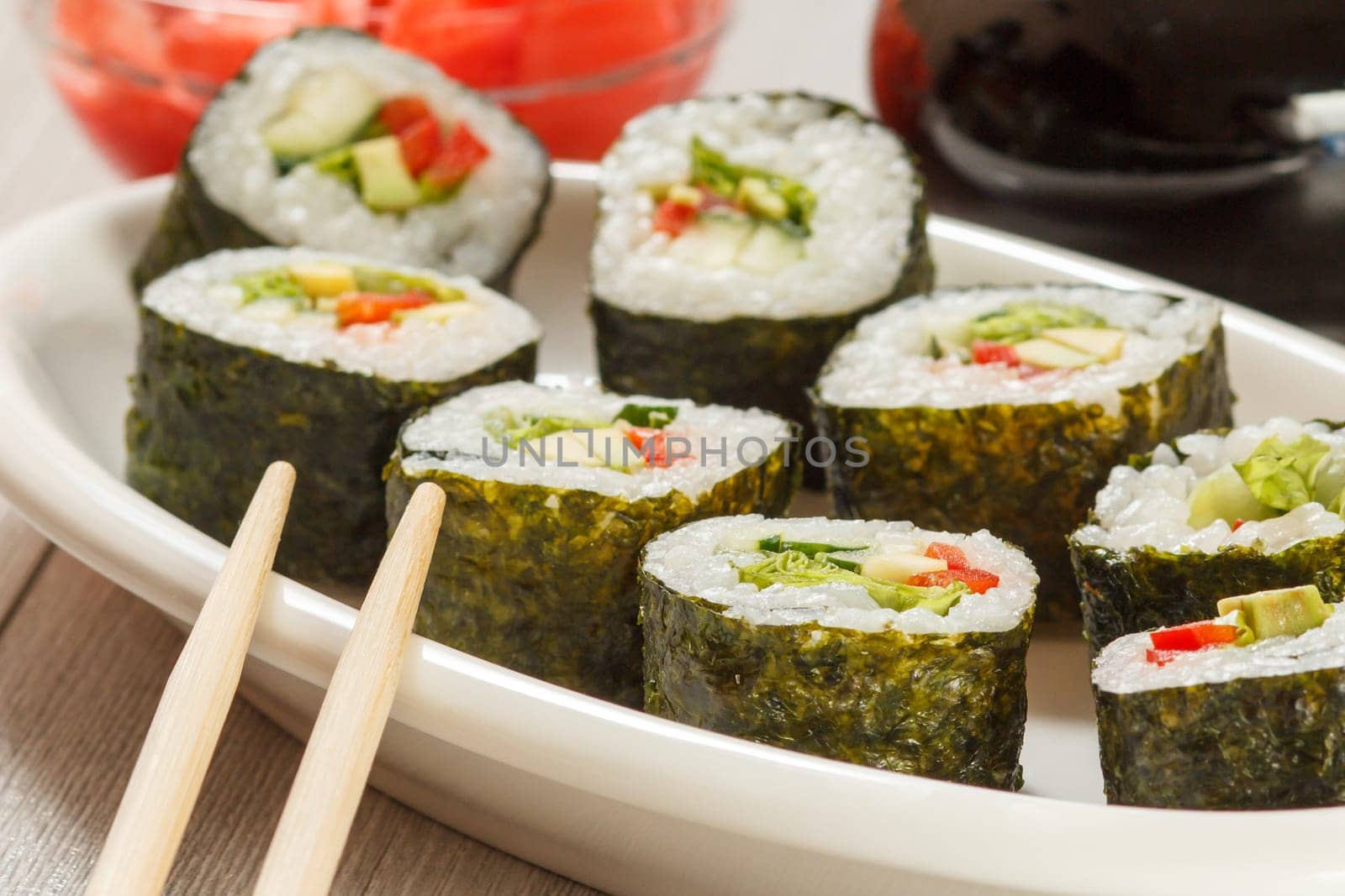 Sushi rolls with rice, pieces of avocado, cucumber, red bell pepper and lettuce leaves on ceramic plate, chopsticks, glass bottle with soy sauce and pickled ginger in the background. Vegetarian food.