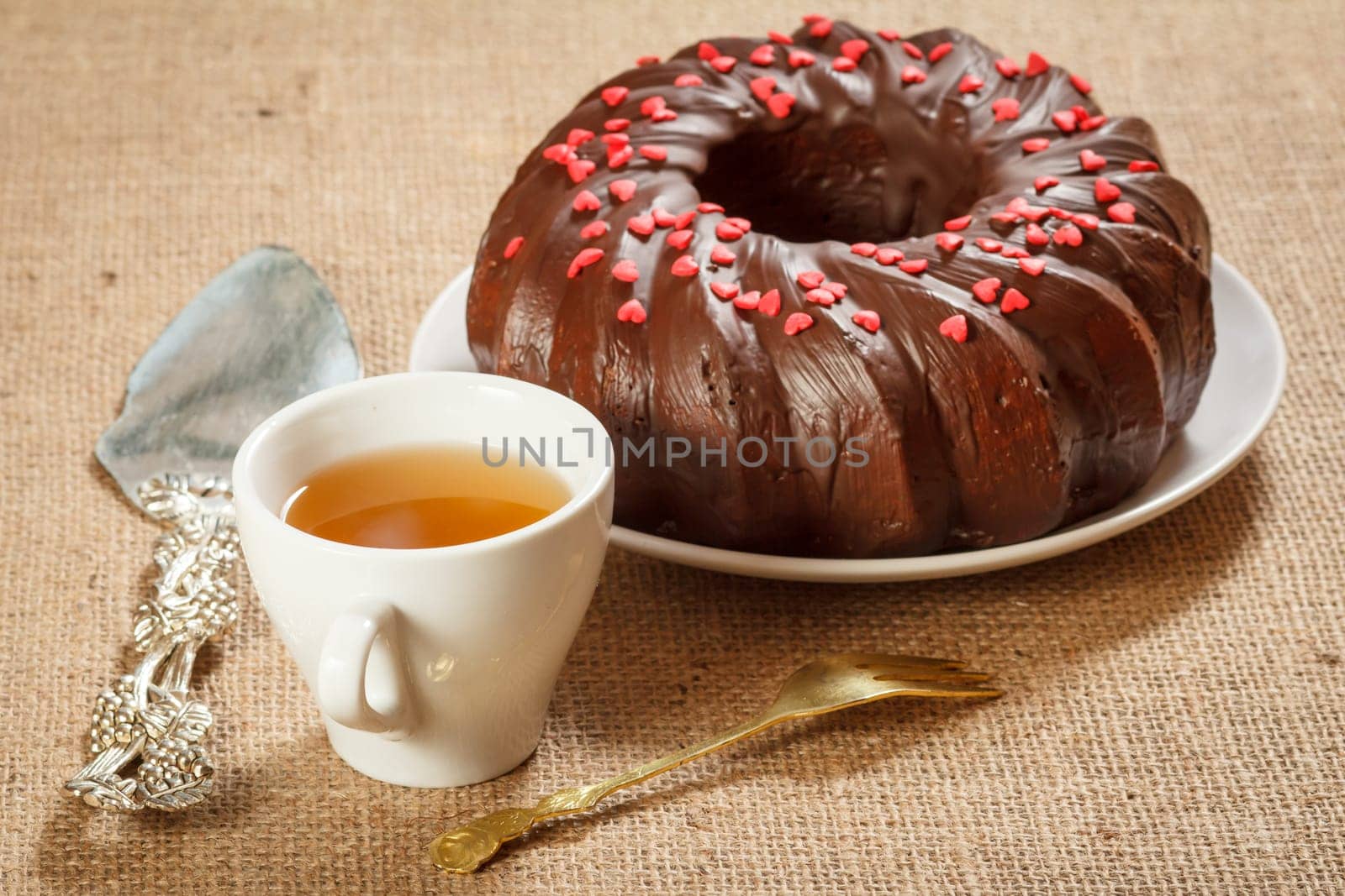 Cup of tea, homemade chocolate cake decorated with small hearts made of caramel on plate, silver cake lifter beside it and fork on table with sackcloth.