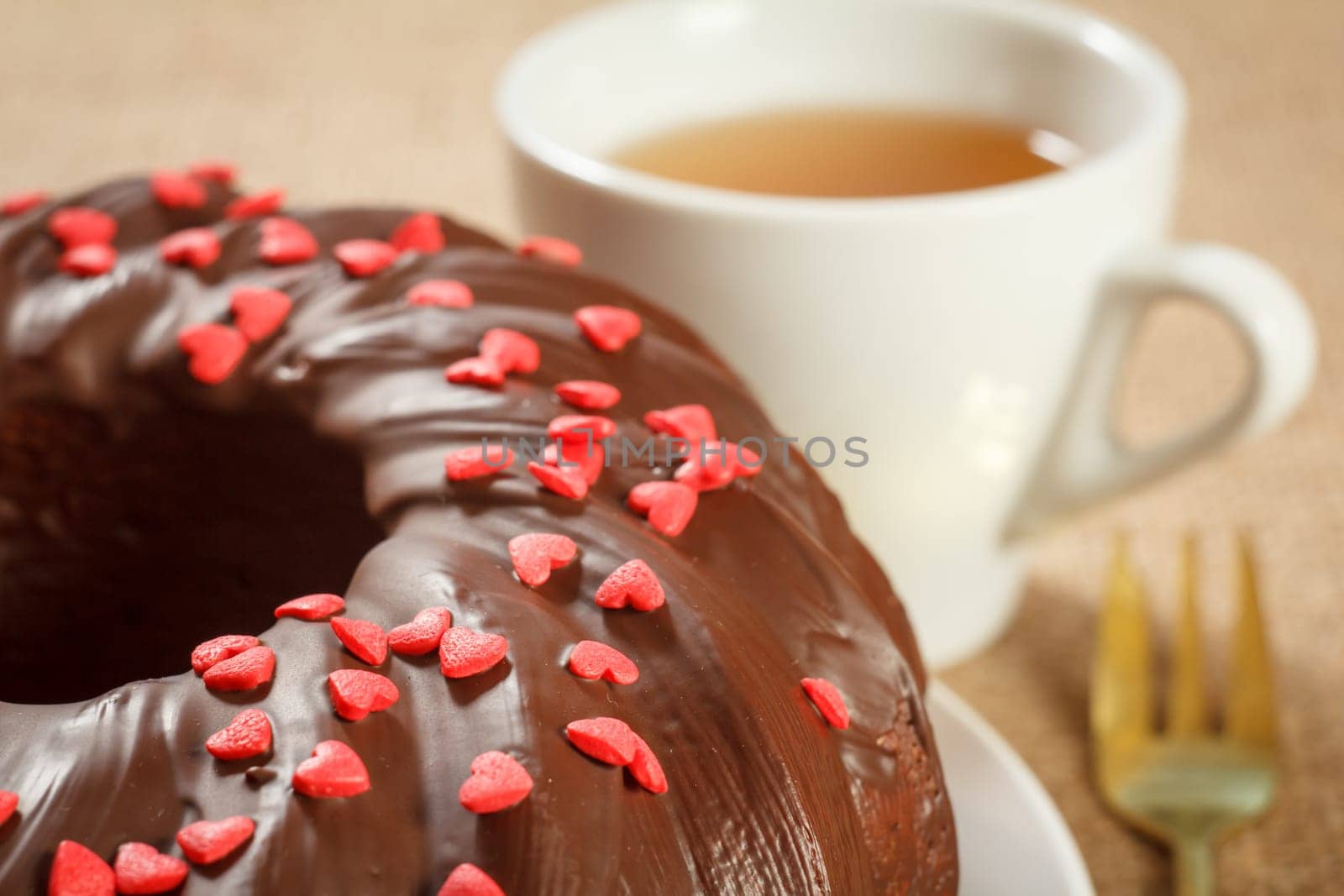 Close-up homemade chocolate cake decorated with small hearts made of caramel on plate and cup of tea on table. Shallow depth of field.