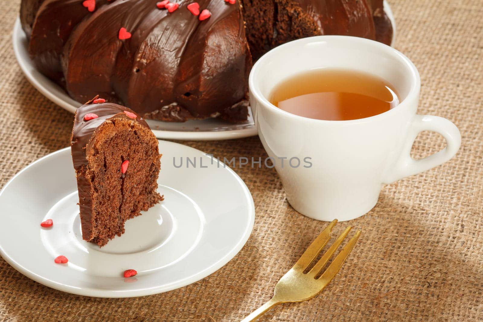 Slice of delicious homemade chocolate cake decorated with small hearts made of caramel on saucer, fork and a cup of tea on table with sackcloth.