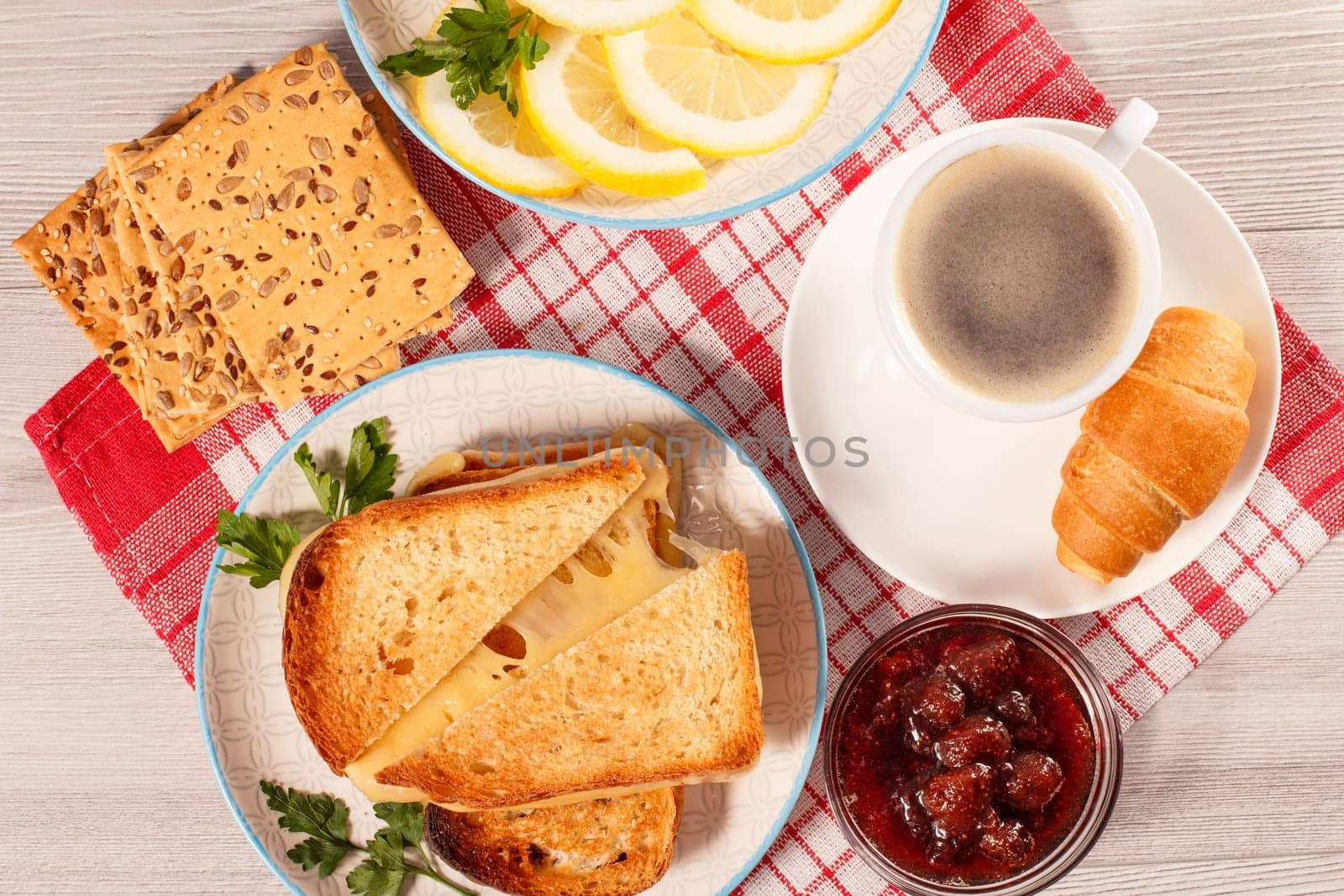 Cup of coffee, toasted slices of bread with cheese and green parsley on white plate, glass bowl with strawberry jam and lemon with red kitchen napkin. Top view. Good food for breakfast.