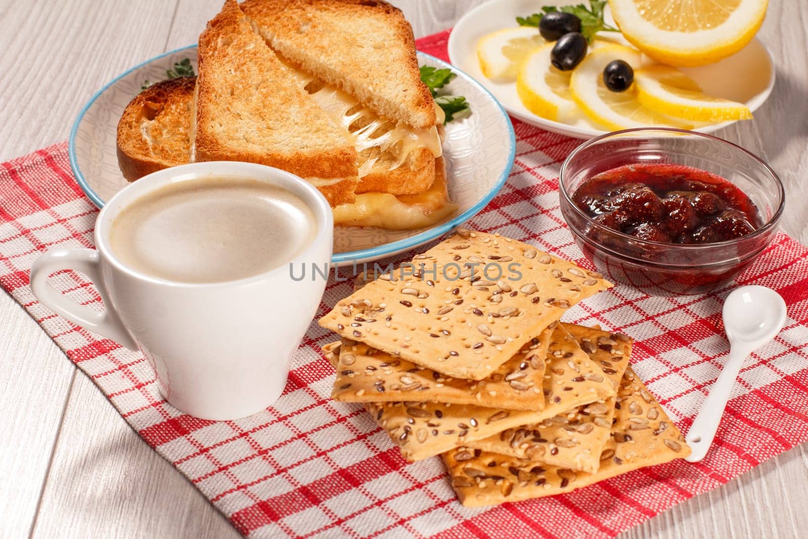 Cup of coffee, toasted slices of bread with cheese and green parsley on white plate, glass bowl with strawberry jam and spoon with red kitchen napkin. Top view. Good food for breakfast.
