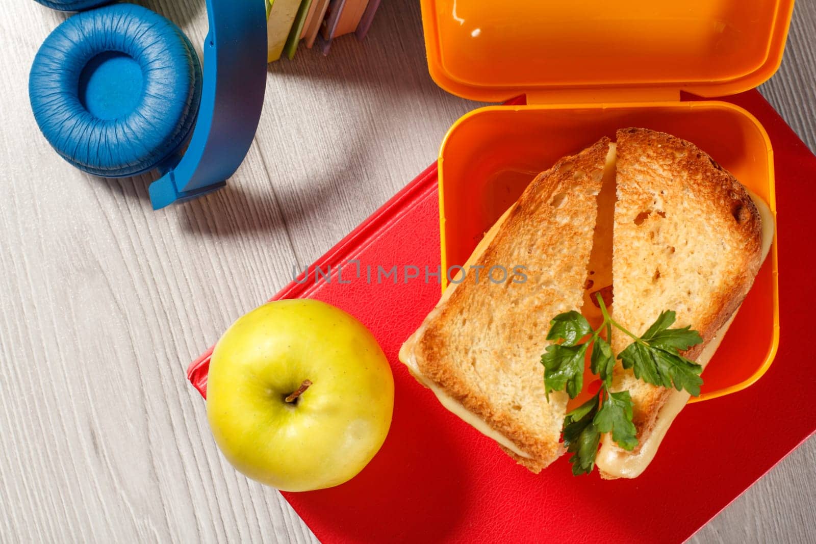 Yellow lunch box with toasted slices of bread, cheese and green parsley, green apple, book and headphones on the background. School breakfast.