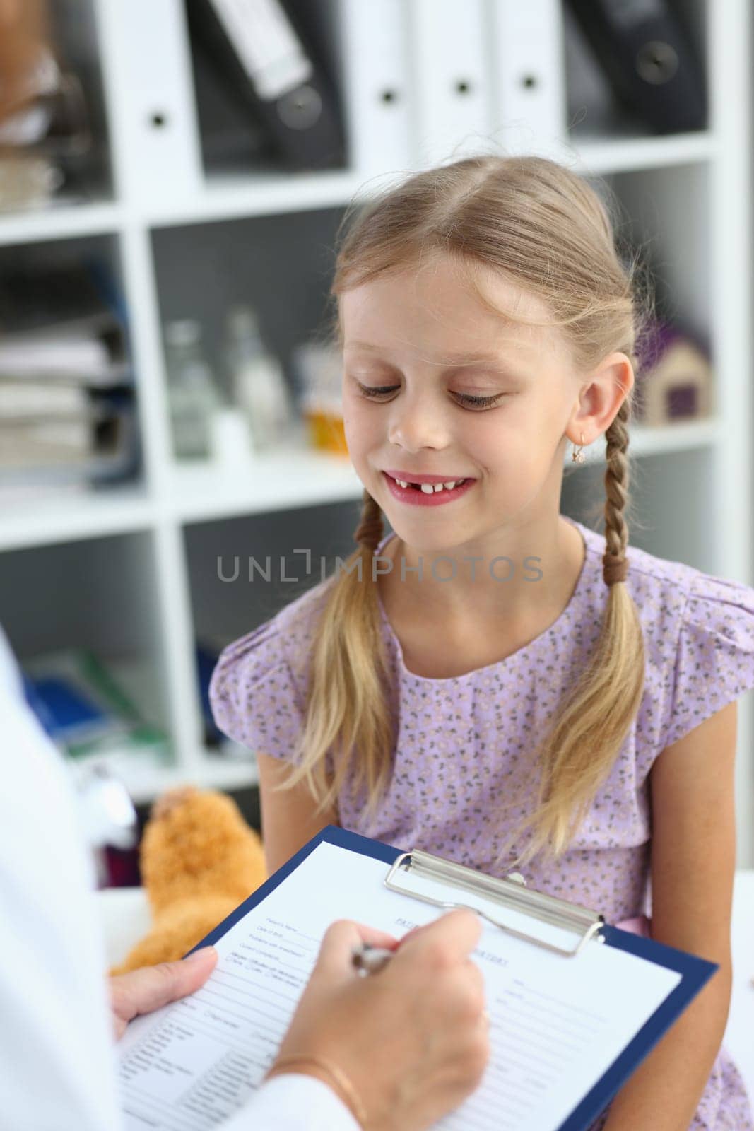 Friendly female pediatrician doctor holding clipboard listening to child girl. Pediatrician talking to child filling out patient form at doctor appointment