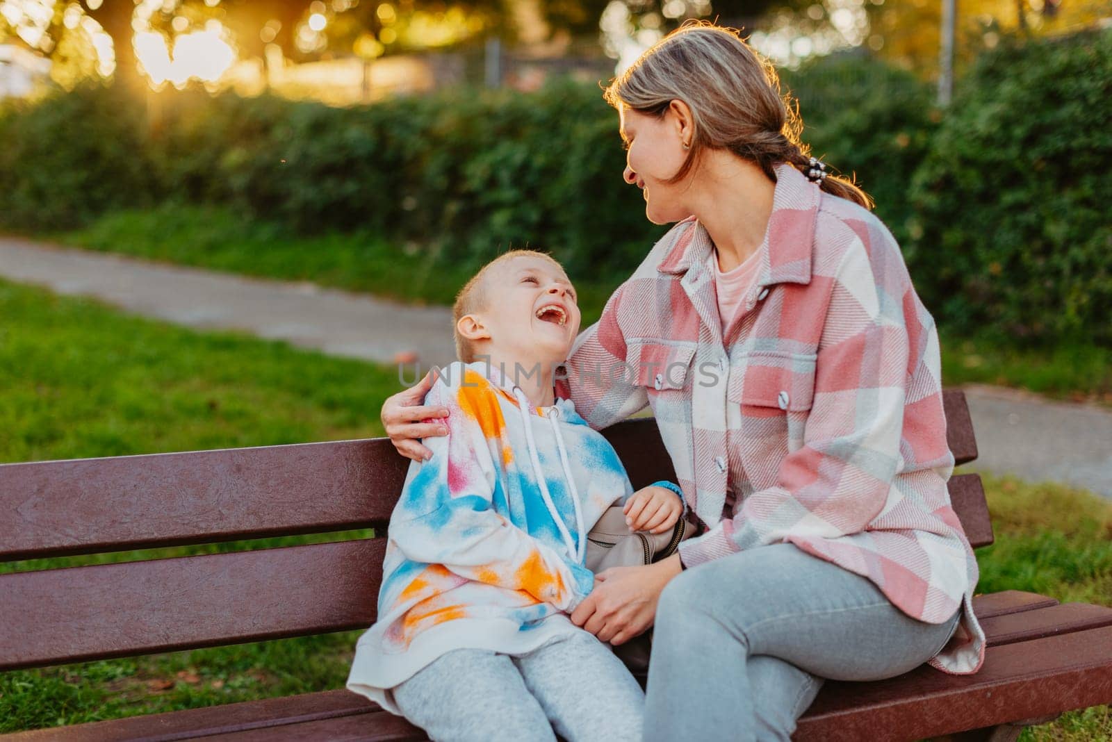 mother and son sit on a park bench in the rays of the setting sun. the concept of a family. Mother's Day. beautiful girl (mother) with a boy (son) in the park in the park are sitting on a bench at sunset by Andrii_Ko