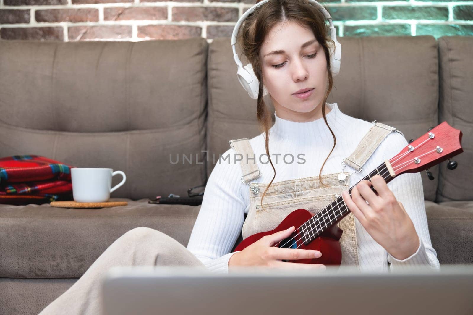 a caucasian girl listens to music through headphones, prepares to play the guitar or ukulele in her house sitting on the floor and looks into a laptop.