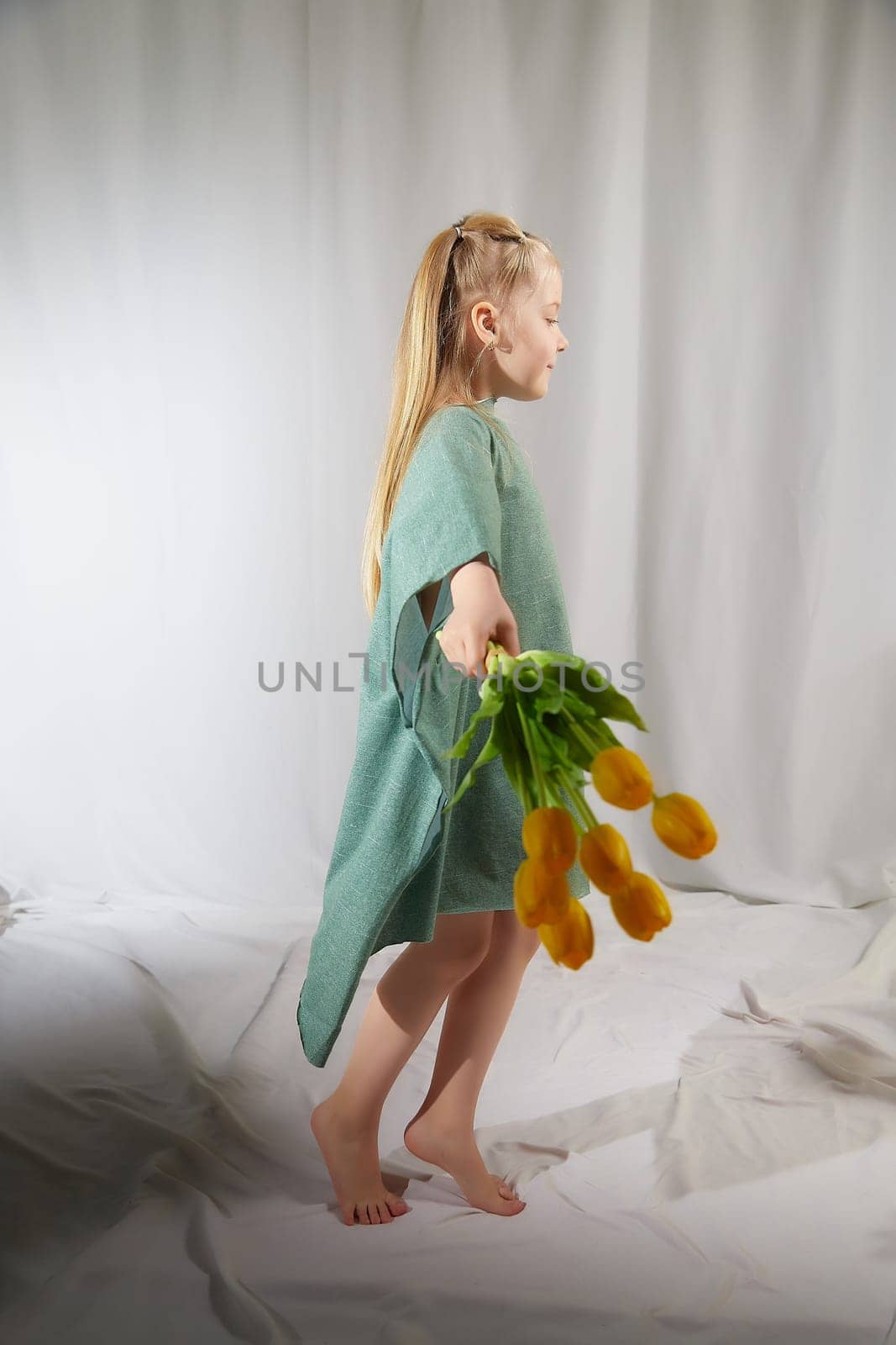 Portrait of a little blonde girl with bouquet of spring yellow flowers on a light background. Child in green dress holding a bouquet of tulips in hands. Spring concept