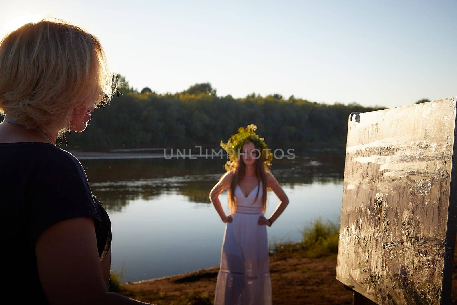 Adult female artist painting picture near water of river or lake in nature. Girl in white sundress and flower wreath posing for holiday of Ivan Kupala in nature at sunset near artist