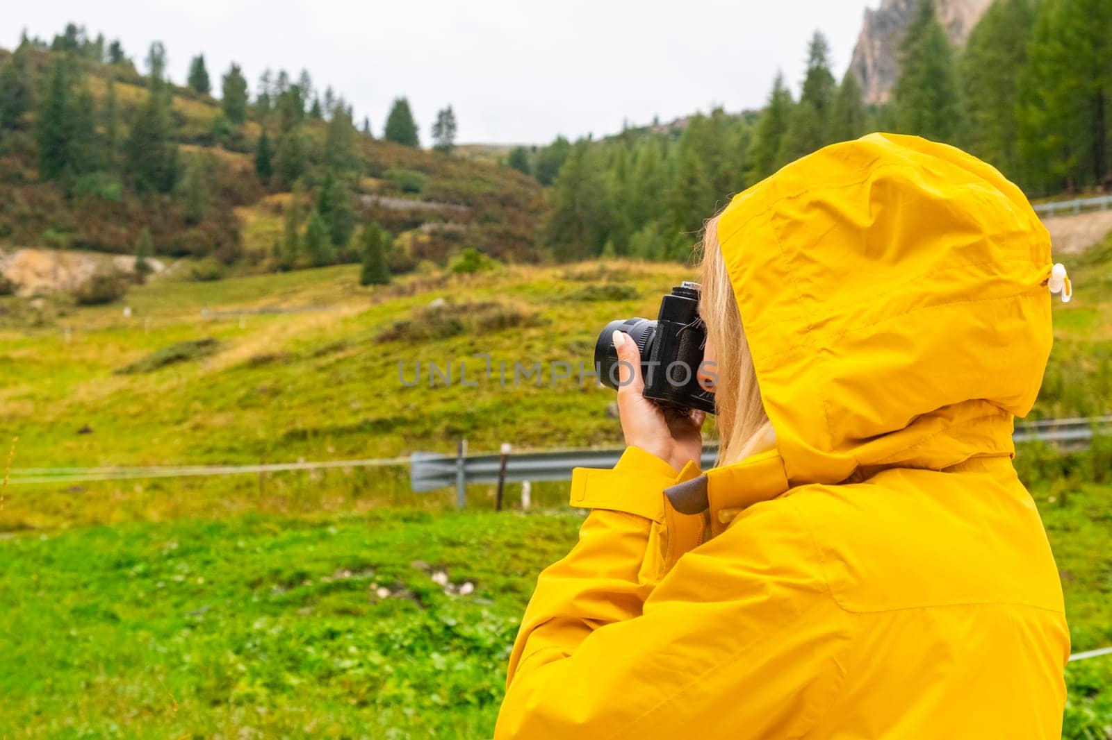 Tourist in a yellow jacket makes photos of meadows of Alpine mountains and forest.