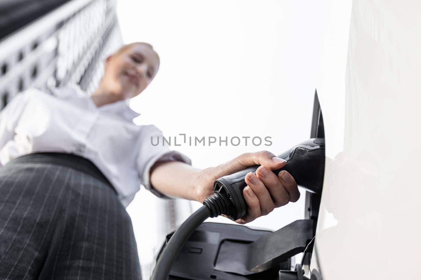 Fisheye view focus hand holding, inserting, plugging ev charger from public charging station for electric vehicle with blurred progressive businesswoman and residential city building in background.