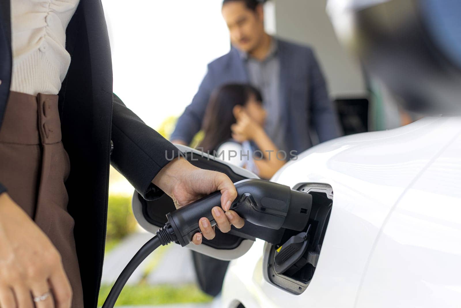 Focus closeup hand charging electric car, insert charger device into electric vehicle as progressive lifestyle concept of alternative green energy technology with blurred family in the background.