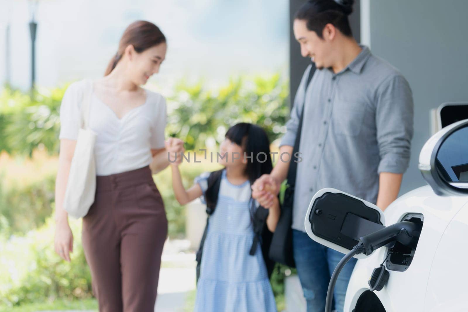 Focus closeup electric vehicle recharging battery from home electric charging station with blurred family in background. Renewable clean energy car for progressive eco awareness lifestyle concept.