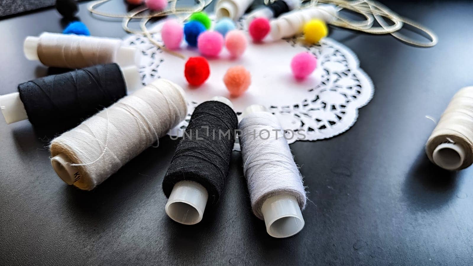 White and black spools of thread with decorative napkin and colored soft fluffy balls on a black background. Black card with abstract frame, texture and sewing threads. Top view. Partial focus by keleny
