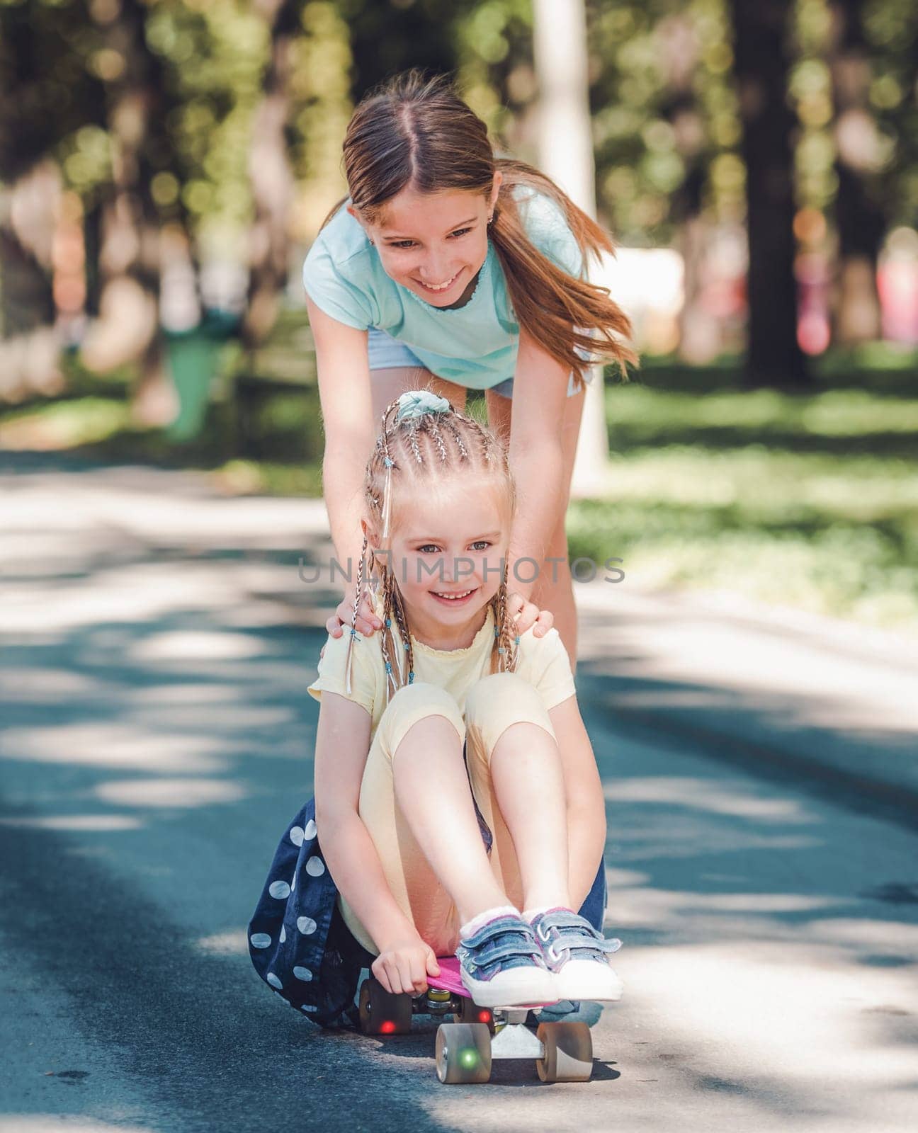 Beautiful teenage girl riding her little blond sister on skateboard in the park.
