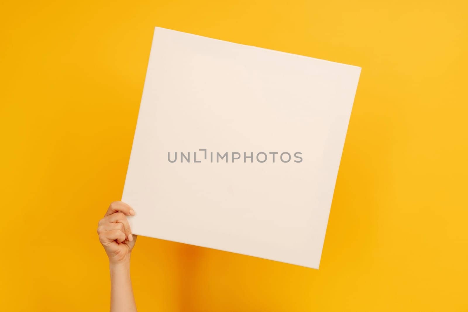 Woman hand holding white canvas paper blank with copyspace on yellow background. Empty sheet banner with place for text. Concept of design and creativity