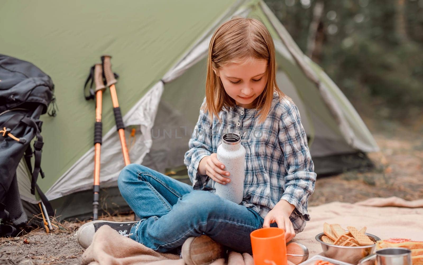 Beautiful preteen girl sitting in the forest in camping and drinking tea from the cup. Cute caucasian child making picnic with tent in the wood