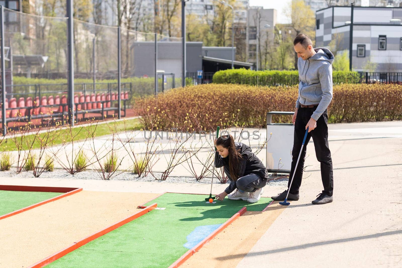 Sharing with golf experience. Cheerful young man teaching his daughter to play mini golf at the day time. Concept of friendly family.