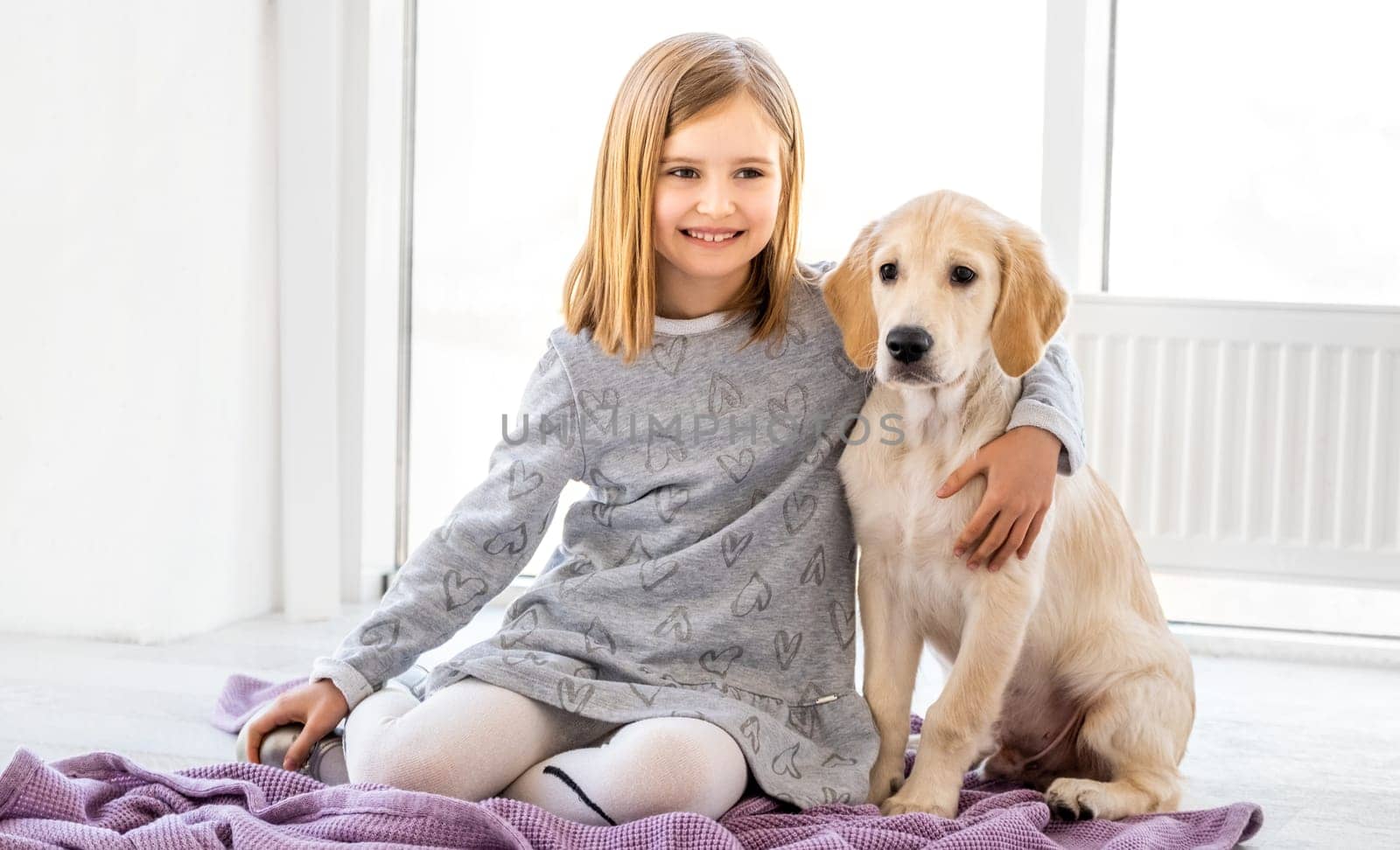 Happy little girl hugging cute dog sitting on floor in light room