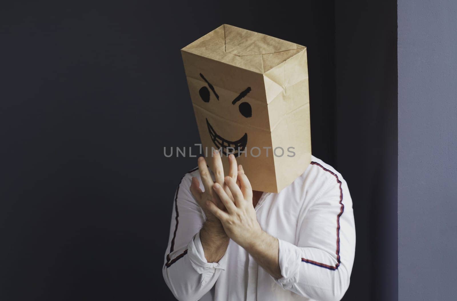 A man in a white shirt with a paper bag with an angry emoticon on his head shows a threatening gesture. Emotions and anger