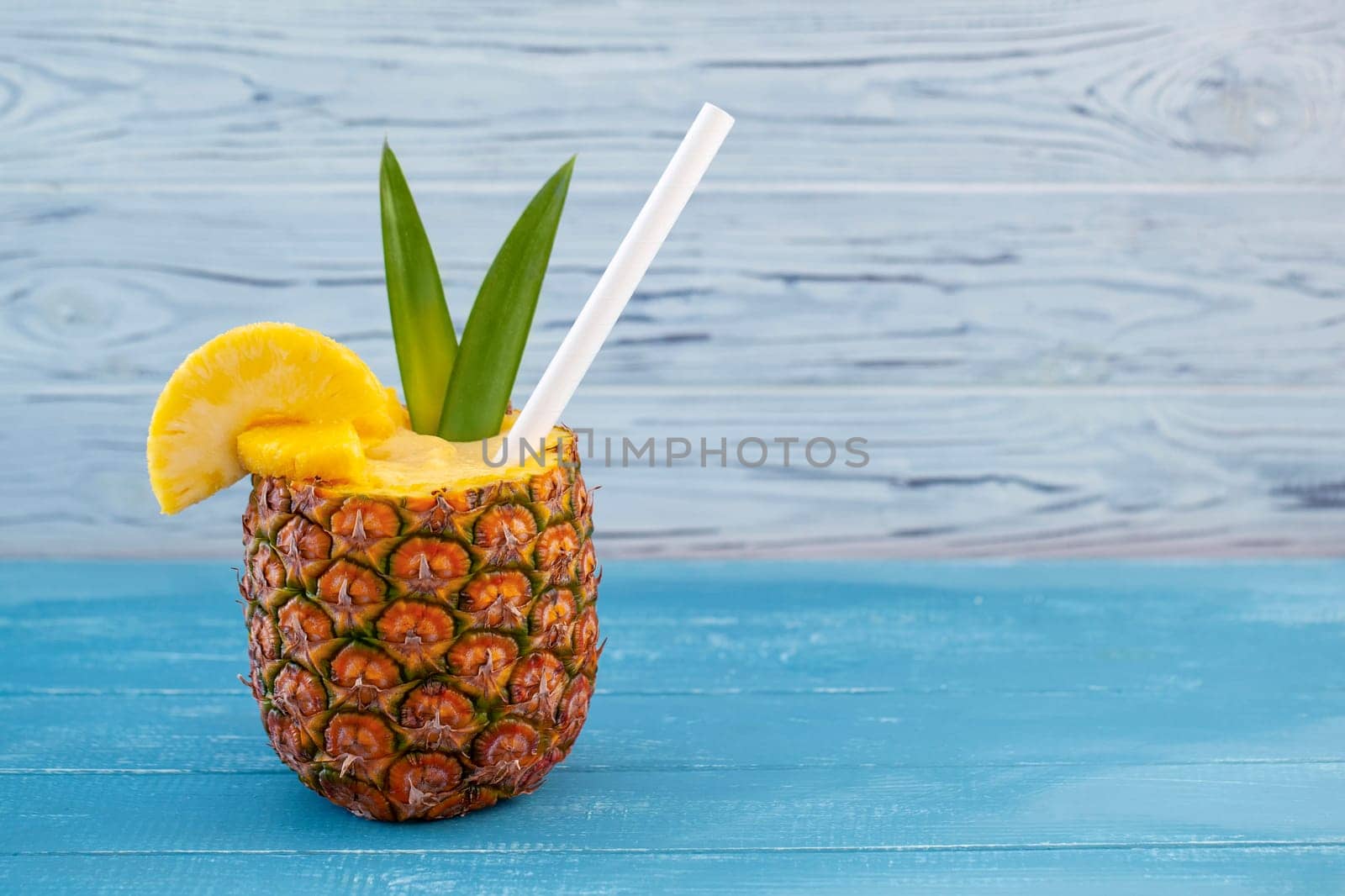 A horizontal photo featuring a pina colada cocktail served in a hollowed-out pineapple with pineapple wedges, a straw, and green leaves on a blue wooden background with copyspace on the right side of the frame