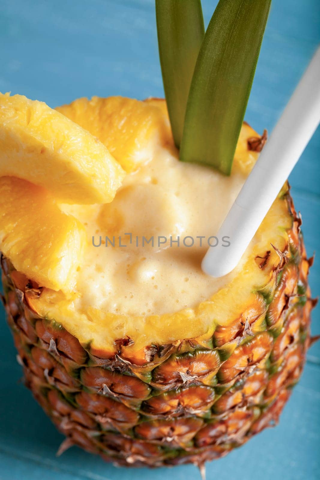 A close-up vertical photo featuring a pina colada cocktail served in a hollowed-out pineapple with pineapple wedges, a straw, and green leaves on a blue wooden background