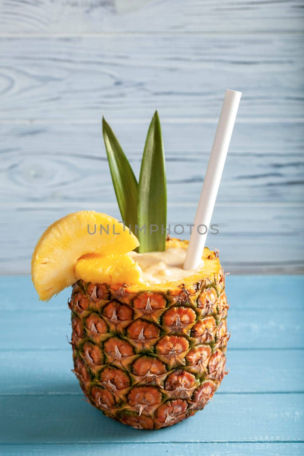 Vertical photo - Pina colada cocktail in pineapple with pineapple slices, straw, two green leaves standing on blue boards