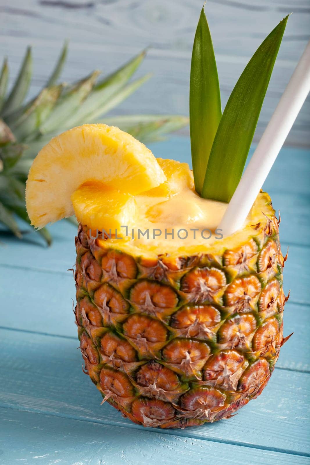 A vertical photo of a pina colada cocktail on a blue wooden background with pineapple pieces, a white straw, two green leaves, and a sliced pineapple crown lying in the blurred background