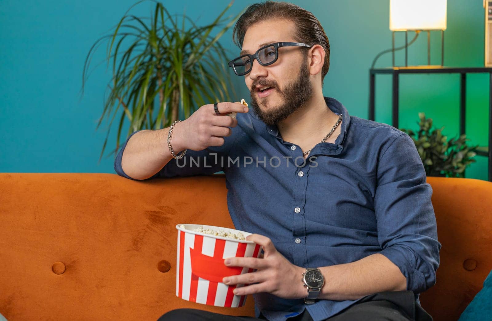 Excited lebanese man in 3D glasses sits on couch eating popcorn snacks, watching interesting TV serial sport game, film online social media movie content at home apartment. Guy enjoying entertainment