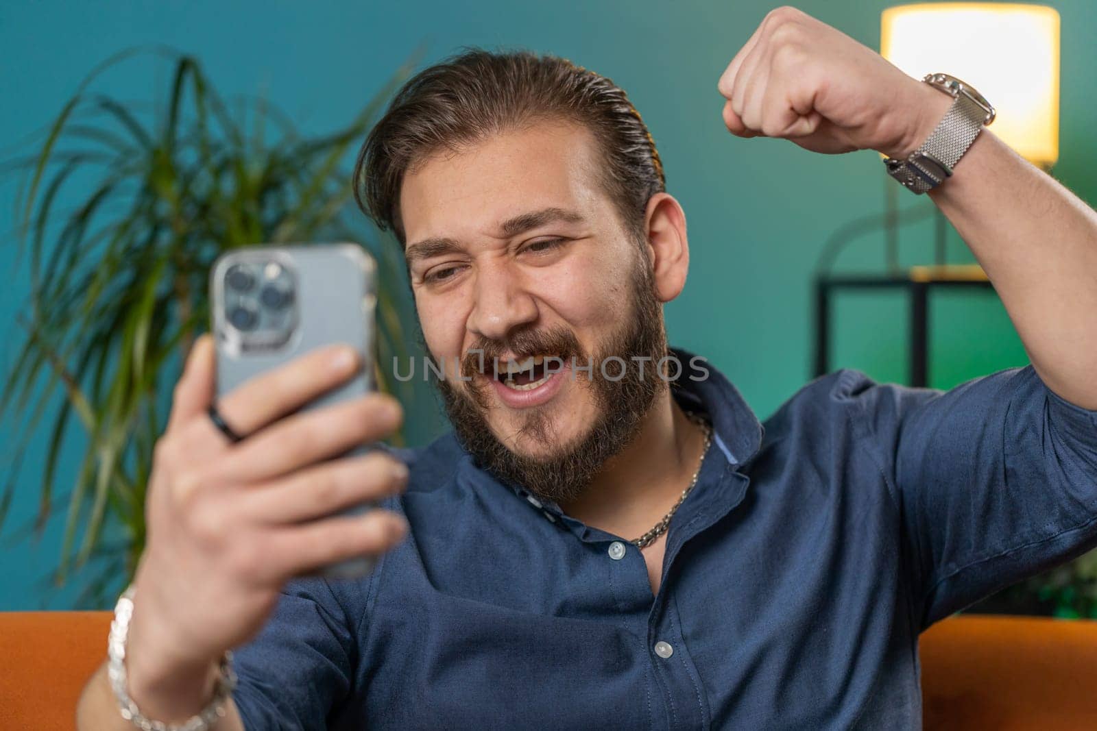 Happy lebanese man use mobile smartphone typing browsing say Wow yes found out great big win good news celebrate lottery jackpot doing winner gesture. Arabian young guy at home in room sitting on sofa