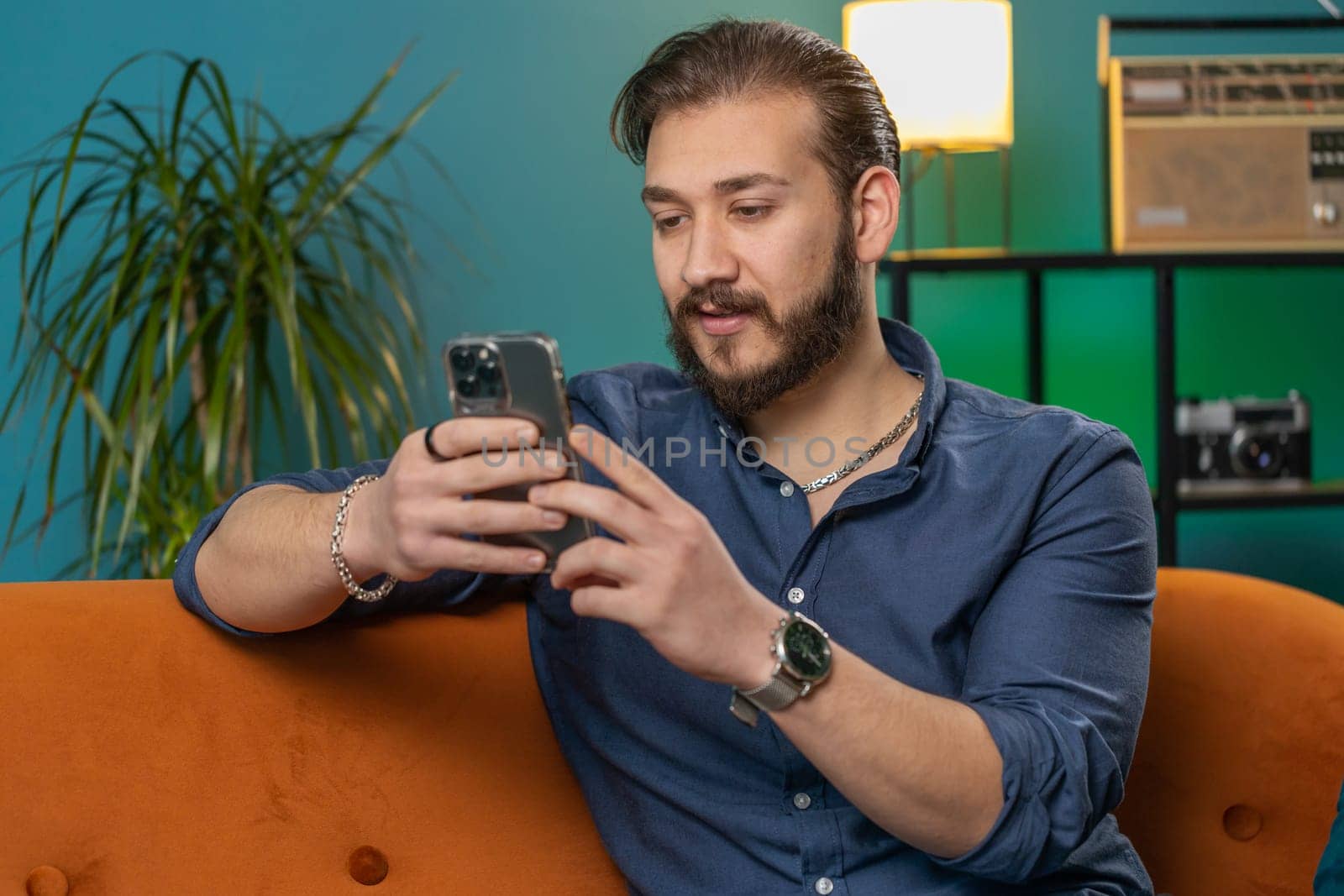 Lebanese man sitting on sofa uses mobile phone smiles at home living room apartment. Arabian guy texting share messages content on smartphone social media applications online, watching relax movie