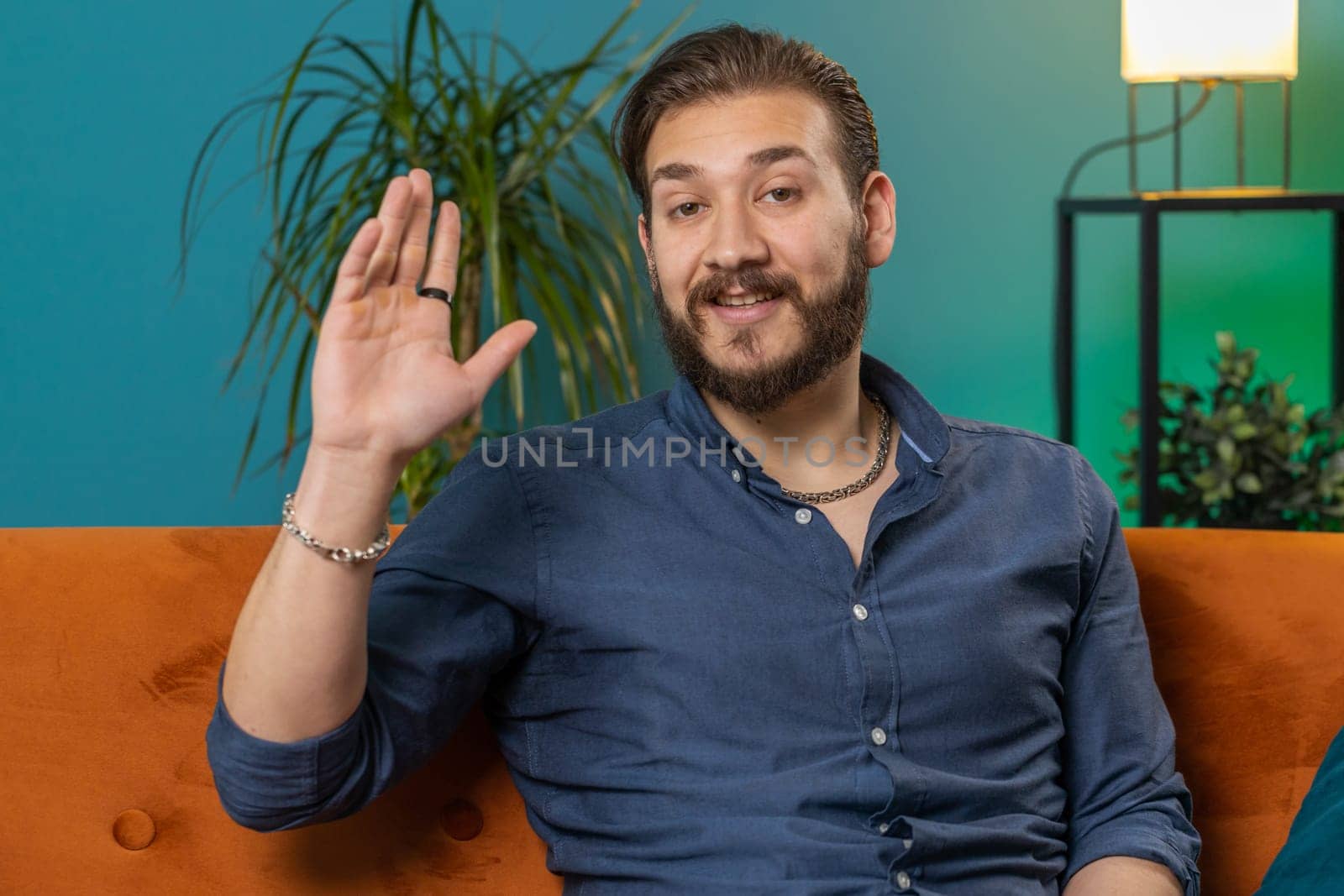Hello. Lebanese man smiling friendly at camera and waving hands gesturing hello, hi, greeting or goodbye, welcoming with hospitable expression at home. Arabian guy sitting on sofa in living room