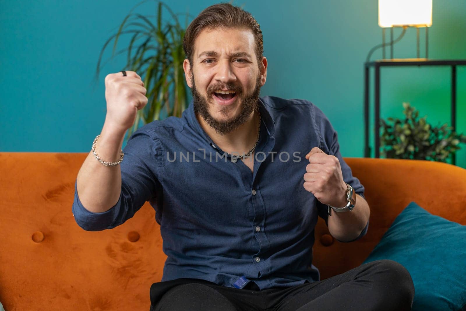 Happy lebanese man shouting, celebrating success, winning, goal achievement good news, lottery luck, victory, watching TV championship at modern home apartment. Arabian guy in room sitting on couch