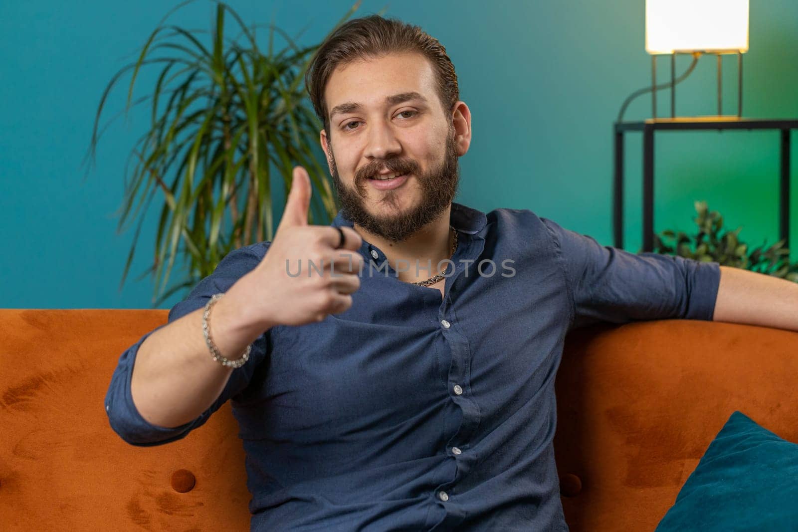 Like. Happy excited lebanese man looking approvingly at camera showing thumbs up, like sign positive something good great news positive feedback. Young arabian guy sitting on couch at home living room
