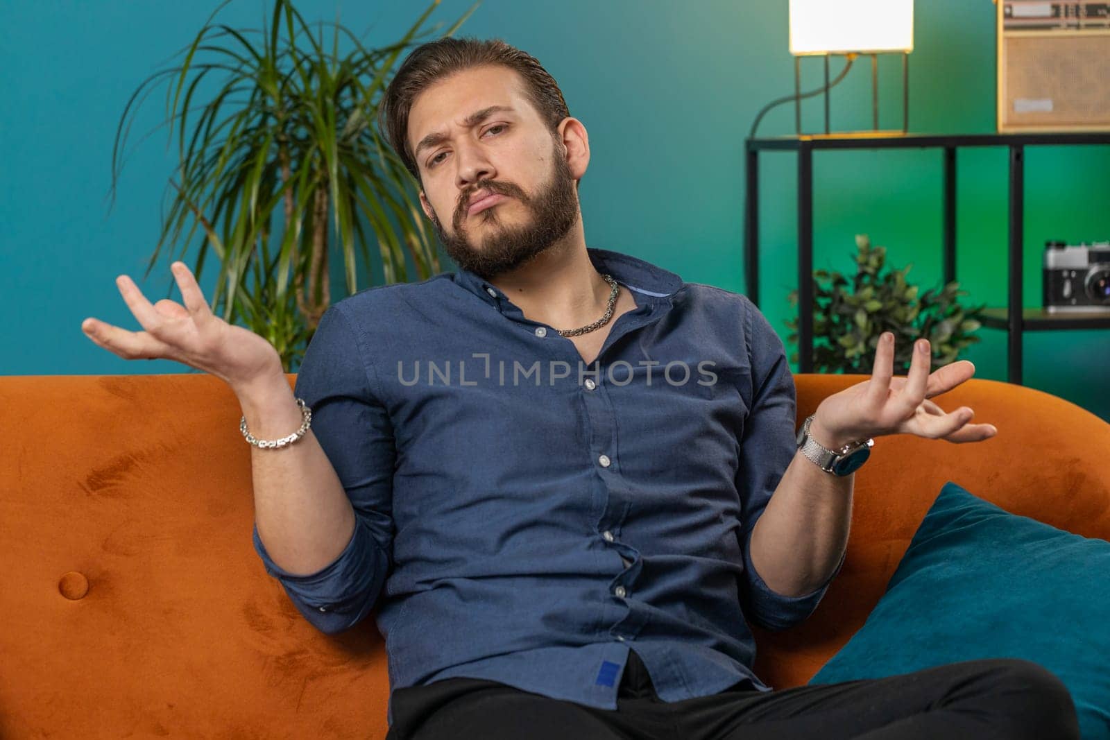 I dont know what to say. Confused lebanese handsome man feeling embarrassed about ambiguous question, having doubts, no idea, being clueless and uncertain at home. Arabian guy sits on sofa in room